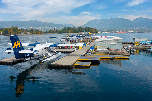 The Harbour Air Launch Pad at Vancouver Harbour Flight Centre, Coal Harbour, Vancouver, Canada. Seaplanes are docked waiting to take passengers out on trips. some people are boarding a seaplane. The Chevron floating fuel barge can be seen further back in the harbour.