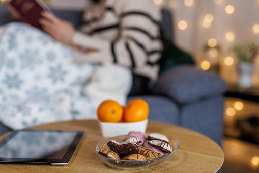 Selective focus shot of unrecognizable young woman lounging on the cozy sofa, under a warm blanket, reading an interesting book. Oranges and cookies served on the coffee table in the foreground.