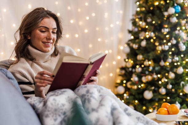 istock Young woman lounging on the sofa, under a warm, cozy blanket, reading a book 1730556574