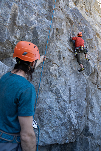 Young man tries to climb a rock while his colleague holds the safety harness