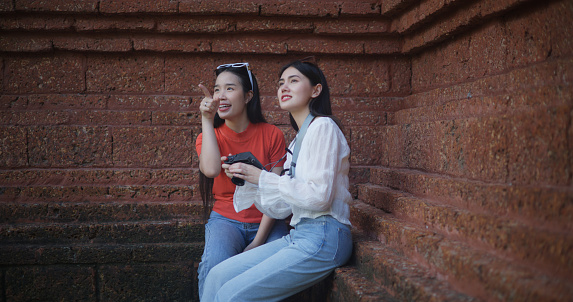 Portrait of Asian women enjoying exploring an ancient temple, sitting together to admire photos on the temple wall. The image embodies travel, people, and lifestyle concepts.