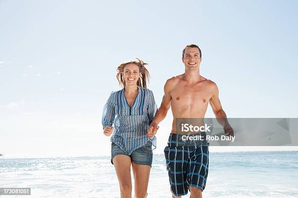 Photo libre de droit de Souriant Couple Courir Sur La Plage Tenant Les Mains banque d'images et plus d'images libres de droit de Hommes