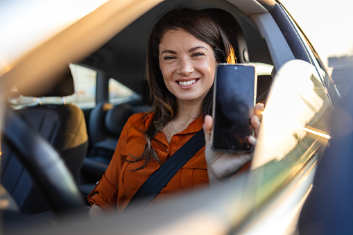 Car Renting App. Happy Female Showing Blank Smartphone While Sitting Inside Of Auto, Smiling Woman Demonstrating Copy Space For Mobile Advertisement Or Website, Mockup