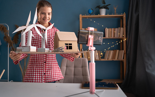 Smiling teen girl recording blog talking about eco project with wind turbines to camera of mobile phone on tripod at home. School influencer filming vlog for her channel. Blogging Concept.