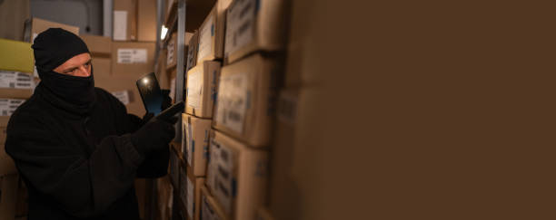istock Thief in a black ski mask stealing boxes at night in a warehouse shines a flashlight in the dark on shelves with goods 1730981017