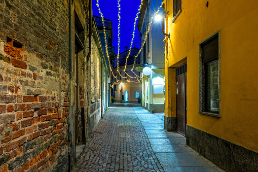 Narrow cobblestone street among old houses illuminated with Christmas lights in evening in Alba, Piedmont, Northern Italy.