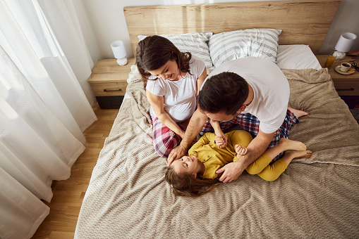 High angle view of happy parents having fun with their small daughter during morning time on a bed in bedroom. Mother is pregnant. Copy space.