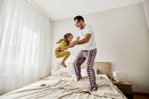Cheerful single father and daughter jumping on a bed.