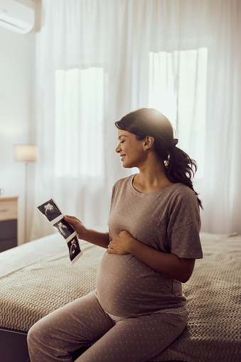 Happy pregnant woman looking at ultrasound of her baby while relaxing on a bed in bedroom.