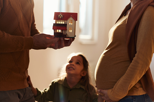 Unrecognizable father holding model home while being with his daughter and pregnant wife in the apartment.