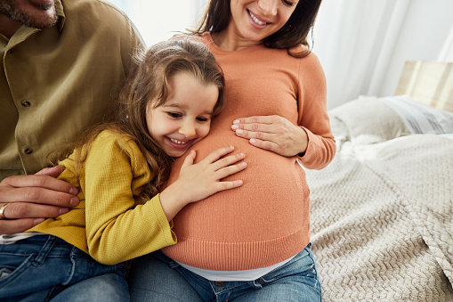 Happy little girl leaning on her mother's pregnant belly in bedroom.