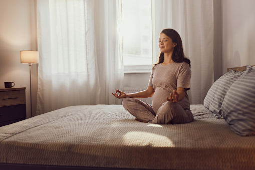 Smiling expecting woman doing Yoga meditation exercises in Lotus position on a bed in the morning. Copy space.