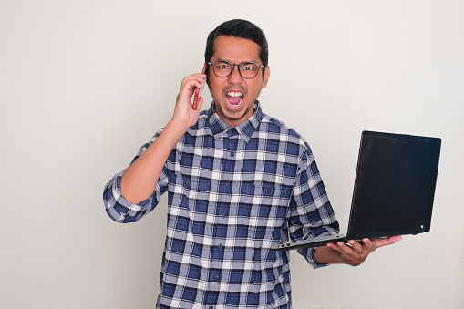 Adult Asian man screaming mad while answering a phone call and holding laptop