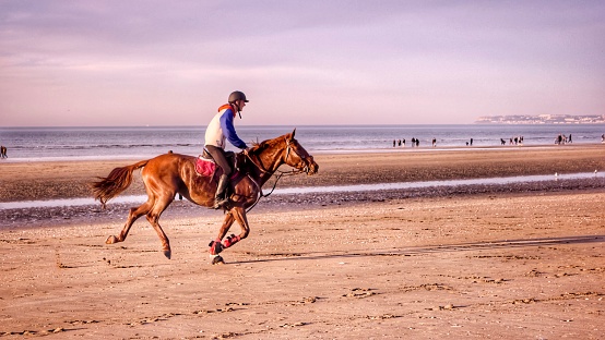 Deauville, France – July 11, 2023: A man wearing a hat and jeans riding a horse along a beach with sparse sand and cloudy skies.