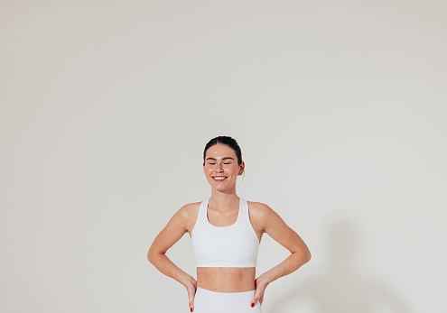 Smiling woman with closed eyes and hands on hips wearing white fitness attire standing at white wall