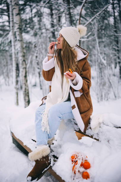 happy smiling young woman eating tangerines in snowy winter forest - snow winter bench park imagens e fotografias de stock