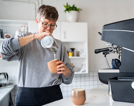 Mid adult woman working in cafe making espresso pouring milk into a cup of coffee