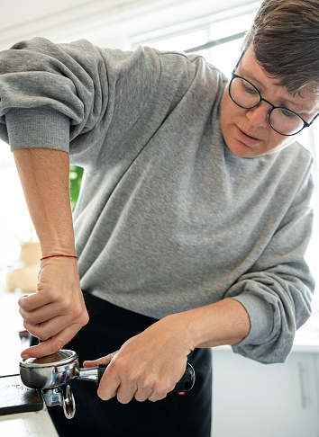 Mid adult woman barista tamping ground coffee with a tamper portafilter while making coffee in cafe kitchen