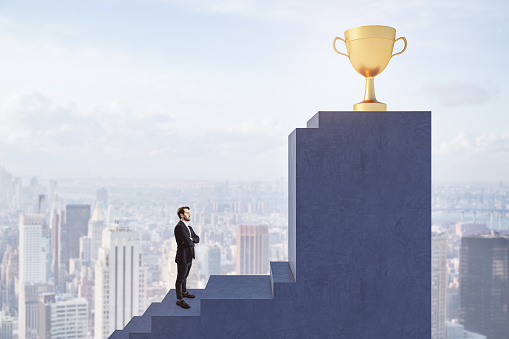 Side view of young european businessman climbing stairs to golden winner's cup with obstacle on blurry city background. Success and career growth concept