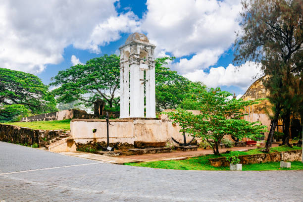 A historical monument in a park-like setting. The monument is a white tower with a dome on top and a clock face on one side. The tower is on a raised platform with steps leading up to it. The platform is surrounded by a low wall with trees and shrubs growing around it. The sky is blue with white clouds and the grass is green. The image is taken from a low angle, looking up at the tower.