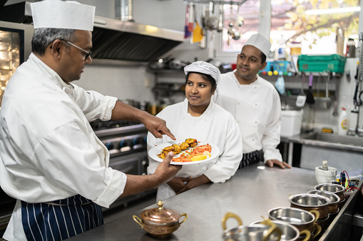 An Indian chef is holding a plate of tandoori chicken and pointing to it as the sous chef's look at him in an industrial catering kitchen