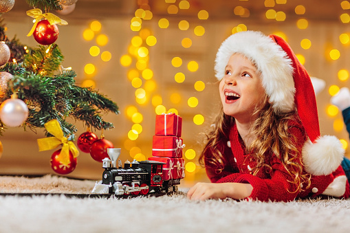 A girl in a Santa hat and a red sweater looks at a festively decorated Christmas tree and waits for miracles. A toy fairy-tale train brings Christmas gifts to children. Background of twinkling lights of garlands.