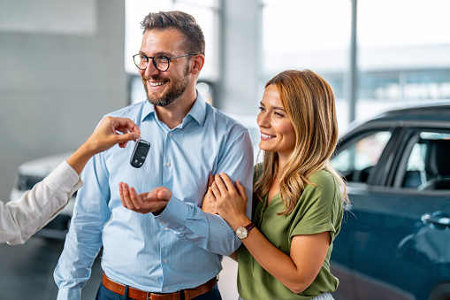 Female dealer giving key of new car to excited young couple in modern auto salon