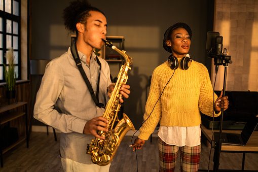 An African-American saxophonist is practising with a female vocalist singing into a microphone in a dark recording studio