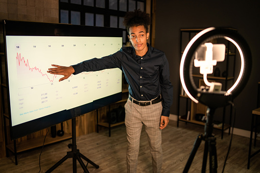 Wearing a dark shirt and smart trousers, an analyst is conducting a financial webinar using a large screen and a cellphone attached to a selfie ring light