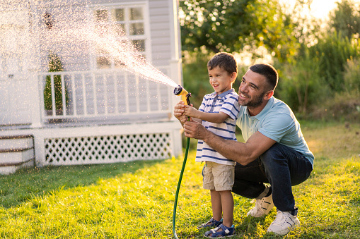 A man is squatting down next to a young boy and holding a garden hose together as they spray water over the grass on a sunny day