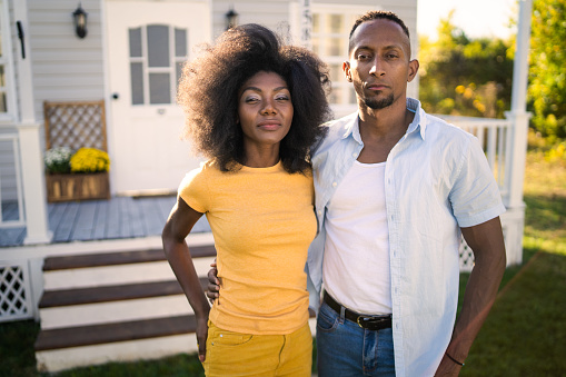 A young African-American couple standing closely together and looking at the camera in front of their house in the garden on a bright sunny day
