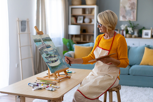 A mature woman sitting on a stool and wearing an apron is painting a picture in her living room