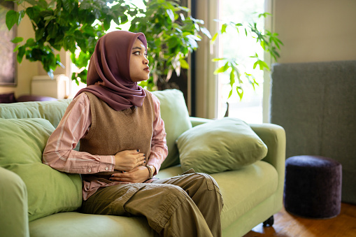 In a modern comfortable room, a young Muslim woman is sitting on a sofa with her hands on her stomach, holding herself as she is in pain, she is gazing into the distance with a worried look on her face