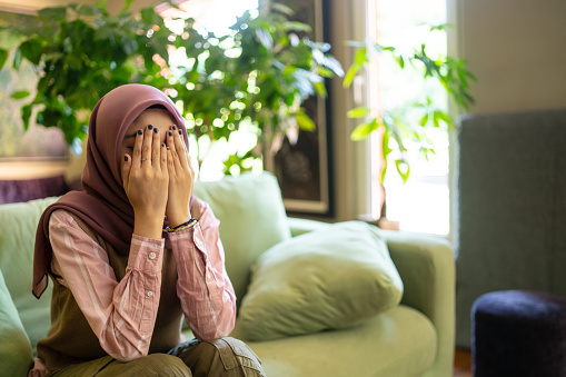 An islamic woman wearing tradition hijab has her hands covering her face as she prays in private to Allah, sitting on her sofa at home