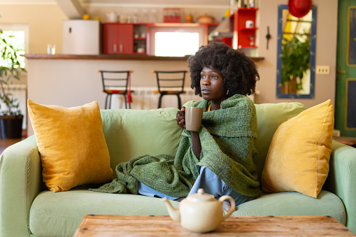 A woman is huddled on her sofa, snuggled into a large blanket around her shoulders as she holds a mug of a hot tea, trying to warm up and feel better while suffering from a cold or flu