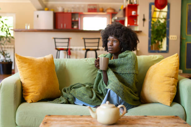 Woman encased in a blanket, with a hot drink A woman is huddled on her sofa, snuggled into a large blanket around her shoulders as she holds a mug of a hot tea, trying to warm up and feel better while suffering from a cold or flu seasonal affective disorder stock pictures, royalty-free photos & images