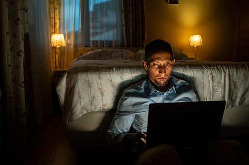 A man sitting up on the floor of his bedroom, leaning against the end of the bed, fully dressed, with his laptop on his knees while he checks his emails late at night