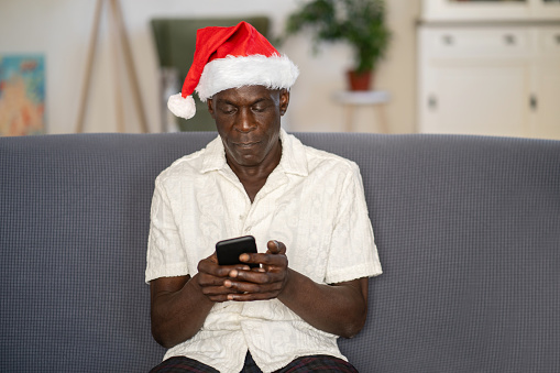 A senior African-American man is sitting on a couch alone, wearing a santa hat for Christmas, he is using his phone to contact his family