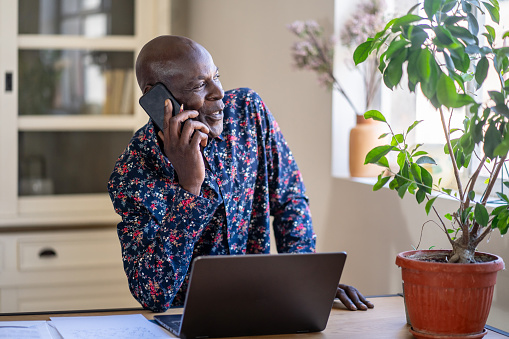 A mature Afro-Caribbeean man is wearing a smart shirt, standing in front of his laptop while he looks out of a window and talks on a smart phone