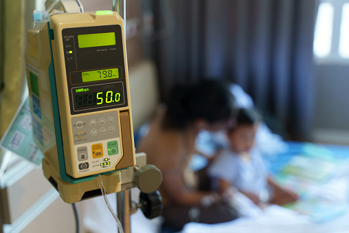 Selective focus on Intravenous IV drip medical machine in a hospital room with child patient and his mother in background.