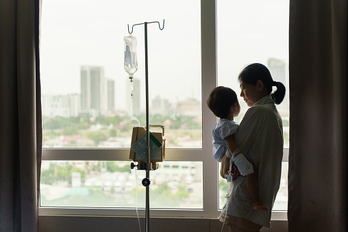Rear view silhouette photo of Asian mother holding and consoling her son with IV drip in hand while recovery in hospital. Adorable Asian kids get sick from virus and hospitalized. Baby health care medical concept.