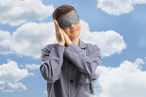 Young man in pajamas wearing a sleeping mask with clouds and sky in the background