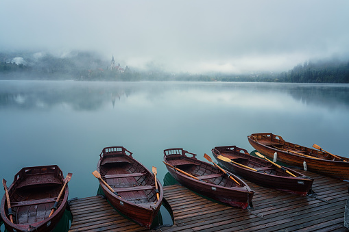 Famous alpine Bled lake (Blejsko jezero) in dense fog, amazing misty landscape, Slovenia. Scenic view of the lake with wooden pleasure boats and island with church, outdoor travel background