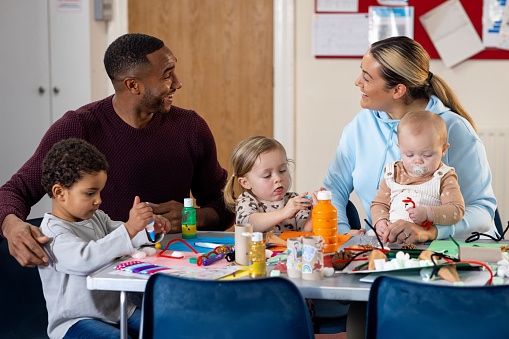 Waist-up shot of a male and a female adult with their children looking after them in a baby class. They are looking over their shoulder while they are being creative. The institute is located in Seghill, Northumberland.
