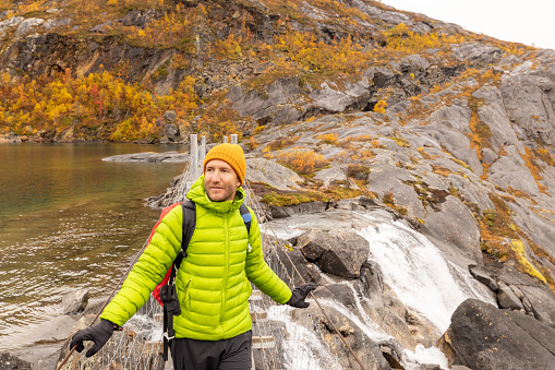 Man hiking in the mountains of Northern Norway in autumn, he crosses a bridge over a waterfall