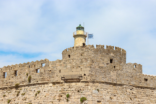 Closeup of Saint Nicholas Fortress round tower with lighthouse and Greece national flag in Rhodes city, Greece