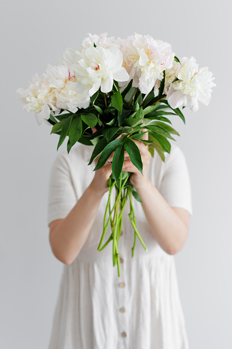 Young woman in dress hiding face behind peony bouquet