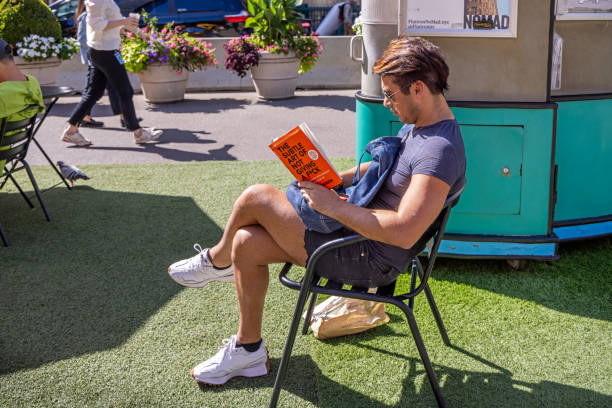 Madison Square, Manhattan, New York, USA - August 23th 2023:  Relaxed young man sitting in a chair on the plastic turf reading a book about not to care