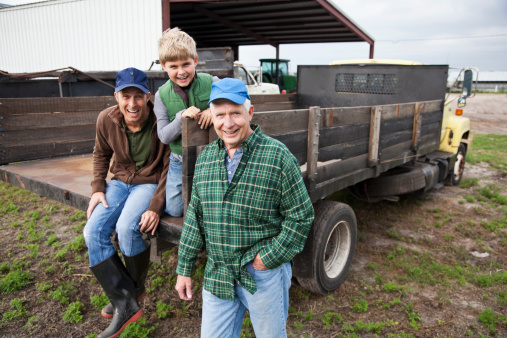 Three generations of men on the back of a truck on the family farm.  The grandfather, a senior man in his 60s, is the focus of the image, standing with his hand in his pocket, smiling at the camera.  His adult son and young grandson are behind him on the truck.  They are wearing jeans, work boots and jackets or flannel shirt.  Everything is brown or green, earthy colors.