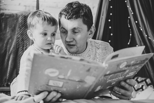 Father reading book for child. Dad hug son enjoying time together. Young family near Christmas tree at home. Merry Christmas and Happy Holidays. Black and white photo. Closeup.